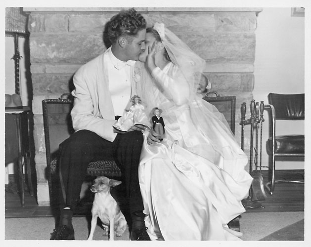 Wally and Rosemarie holding doll replicas with Rosie's dog, Pudgy, 
in front of fireplace in living room